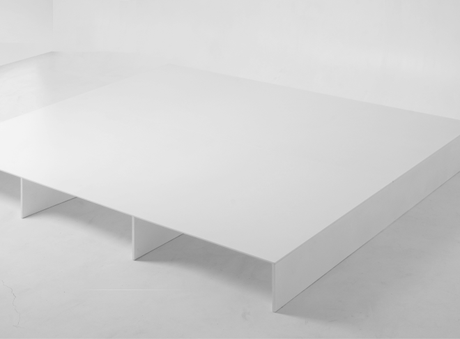 A minimalist white bed platform with a low, flat surface and four vertical support panels underneath. The frame has no headboard.