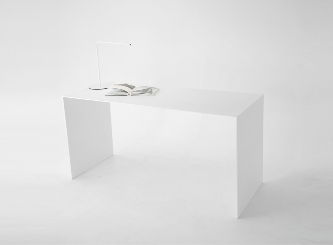 A wide shot of a minimalist U shape white desk. An open book is placed next to the lamp, lying flat on the desk's surface.